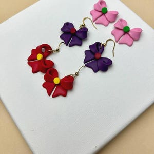 Op de afbeelding: Handgemaakte strikvormige oorbellen in rood, paars en roze. De oorbellen hebben goudkleurige haken en kleine, contrasterende middenaccenten. De strikken worden gepresenteerd op een wit oppervlak.