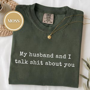 May include: A moss green short-sleeve t-shirt featuring the white printed text My husband and I talk shit about you. The shirt has a Comfort Colors brand label at the inner neckline and is displayed with a gold sunburst pendant necklace.