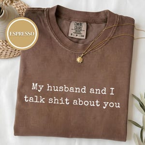 May include: A brown short-sleeved t-shirt with the white text "My husband and I talk shit about you" printed on the front. The shirt features a Comfort Colors brand label at the inner neckline and is displayed with a gold sunburst pendant necklace.