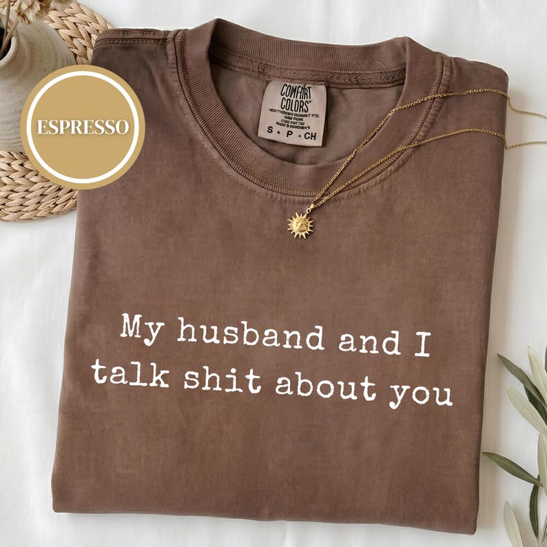 May include: A brown short-sleeved t-shirt with the white text "My husband and I talk shit about you" printed on the front. The shirt features a Comfort Colors brand label at the inner neckline and is displayed with a gold sunburst pendant necklace.