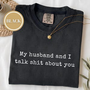May include: A black short-sleeved t-shirt with a crew neckline featuring the white printed text "My husband and I talk shit about you" across the chest. The shirt has a Comfort Colors brand label inside the collar and a gold sunburst pendant necklace.