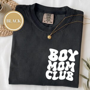 Puede incluir: Una camiseta negra de manga corta con cuello redondo que presenta el texto blanco BOY MOM CLUB en una fuente ondulada y redondeada en la parte inferior derecha. La prenda incluye una etiqueta de la marca Comfort Colors y un collar con sol.