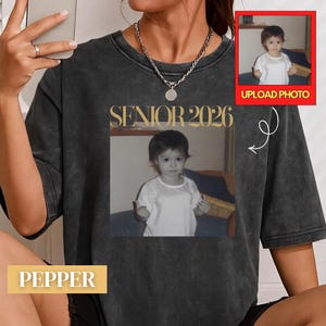 Puede incluir: Una camiseta gris carbón con lavado ácido que presenta una impresión fotográfica personalizada de un niño pequeño con camisa blanca. El texto SENIOR 2026 está impreso en una fuente serif dorada sobre la foto. Un recuadro muestra UPLOAD PHOTO.