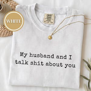 May include: A white short-sleeved t-shirt featuring the black printed text My husband and I talk shit about you. The shirt has a Comfort Colors brand label at the inner neckline and is paired with a gold-toned sunburst pendant necklace.