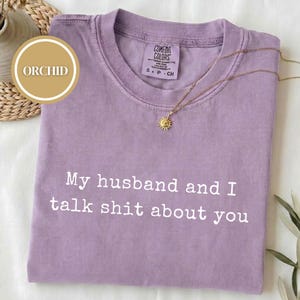 May include: An orchid-colored short-sleeve t-shirt with a crew neckline. The shirt features the white printed text "My husband and I talk shit about you" across the chest. A gold sun-shaped pendant necklace rests on the fabric near the collar.