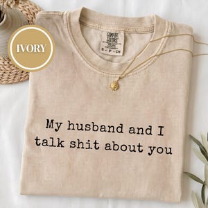 May include: An ivory short-sleeve t-shirt with a crew neckline and a Comfort Colors brand label. The shirt features the black printed text "My husband and I talk shit about you" centered on the chest. A gold sun-shaped pendant necklace is included.