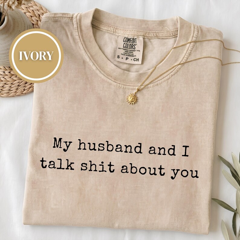 May include: An ivory short-sleeve t-shirt with a crew neckline and a Comfort Colors brand label. The shirt features the black printed text "My husband and I talk shit about you" centered on the chest. A gold sun-shaped pendant necklace is included.
