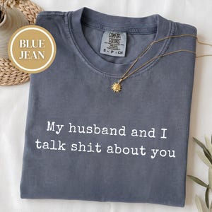 May include: A blue jean colored t-shirt with a crew neckline and a Comfort Colors brand label at the collar. The shirt features the white text "My husband and I talk shit about you" printed across the chest. A gold sunburst pendant necklace rests on the fabric.
