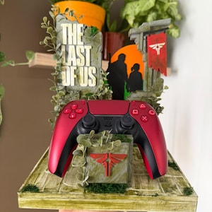 Peut inclure: Une manette de jeu vidéo rouge et noire posée sur un support thématique. Le support présente une base en pierre avec un logo Firefly rouge, un fond avec le texte The Last of Us, une silhouette de deux personnages et une bannière rouge.