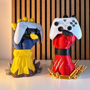 Può includere: Due supporti per controller a forma di mani stilizzate. La mano sinistra è blu e gialla e sostiene un controller PlayStation bianco. La mano destra è rossa con una fascia nera, sostiene un controller Xbox bianco ed è circondata da bossoli dorati.