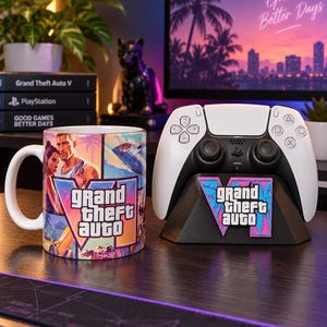 Puede incluir: Una taza de cerámica blanca con un estampado gráfico colorido de Grand Theft Auto VI y un mando de juego inalámbrico blanco sobre un soporte negro con un emblema del logotipo de Grand Theft Auto VI a juego en la parte frontal de la base.