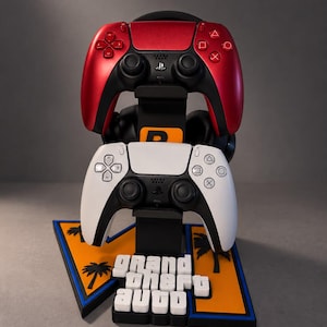 Può includere: Un supporto nero per controller di gioco con un controller rosso metallizzato sopra e uno bianco sotto. La base mostra il logo arancione e blu di Grand Theft Auto con sagome di palme. Un paio di cuffie nere poggia dietro i controller.