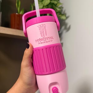 Pode incluir: Um copo térmico rosa claro e rosa escuro com o logótipo Monster Energy em relevo na parte superior. O recipiente inclui uma pega rosa escura, uma palhinha de plástico transparente e uma pequena torneira situada perto da base.