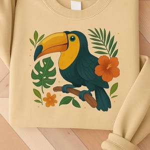 May include: A light yellow sweatshirt featuring a toucan illustration. The toucan has a large yellow and orange beak, with teal feathers and is perched on a brown branch. The design includes green leaves and orange flowers, creating a tropical theme.