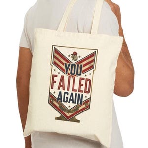 May include: A cream-coloured canvas tote bag featuring a graphic design with red, white, and blue stripes. The text reads "YOU FAILED AGAIN" in bold letters, with a small eagle icon wearing a red hat and the phrase "STAND WITH TRUMP" at the bottom.