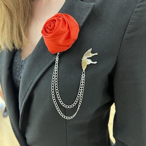 May include: A red fabric rose brooch with a silver chain and a butterfly-wing-shaped pin, attached to a black blazer. The rose is a bright red, and the chain hangs in two strands.