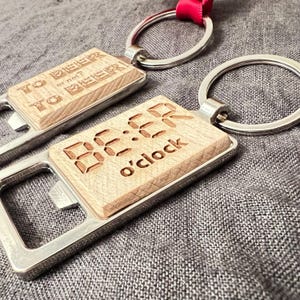 以下が含まれることがあります： 木製のアクセントが付いた2つの栓抜きキーホルダー。1つは「TO BEER or not BEER？」と書かれており、もう1つは「BE:ER o'clock」と書かれています。どちらも銀色の金属製の栓抜きとキーリングが付いています。
