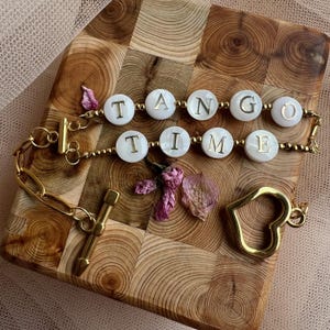 May include: A gold chain bracelet with white shell beads that spell out "TANGO TIME". The bracelet has a heart charm and a toggle clasp.