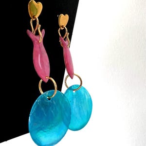 Puede incluir: Un par de pendientes colgantes con tachuelas doradas en forma de corazón. Los pendientes presentan elementos rosas en forma de lágrima conectados a grandes colgantes ovalados de color turquesa. Aros y detalles dorados.