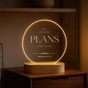 Puede incluir: Lámpara LED circular con las palabras "PLANS I HAVE FOR YOU" y la referencia "JEREMIAH 29:11". La lámpara, con base de madera, está sobre una mesita de noche de madera oscura. La luz emite un brillo cálido.