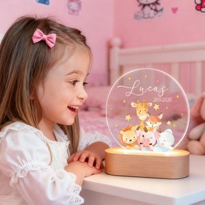 Puede incluir: Una luz de noche personalizada con una base de madera y un panel acrílico circular transparente. El panel presenta el nombre "Lucas" y la fecha "20.5.2026", junto con ilustraciones de una jirafa, un zorro, un león, un cerdo y un elefante. La luz emite un brillo cálido.