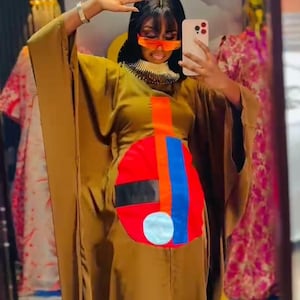 Op de afbeelding: Een lange, vloeiende bruine jurk met een kleurrijk abstract ontwerp op de voorkant. Het ontwerp bevat rode, blauwe en oranje vormen. De jurk heeft wijde mouwen en wordt gedragen met een statement ketting en oranje getinte glazen. De afbeelding is genomen voor een spiegel.