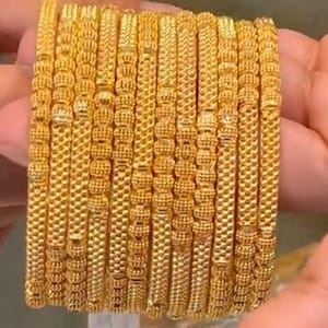 May include: A set of gold bangles. The bangles feature detailed textured patterns, with alternating sections of beaded and woven designs. The gold jewellery is shown in a close-up, highlighting the craftsmanship.