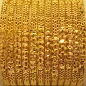 Könnte beinhalten: Ein Satz goldener Armreifen mit aufwendigen Perlen- und strukturierten Designs, präsentiert in einer weißen Box. Die Armreifen sind in einem hellen, glänzenden Goldton gehalten und reflektieren das Licht. Der Schmuck wird auf einem weichen, cremefarbenen Kissen präsentiert.