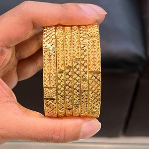 Może przedstawiać: Zestaw czterech złotych bransoletek typu bangle z powtarzającym się geometrycznym wzorem kratki. Każda bransoletka ma polerowane wykończenie ze skomplikowanymi detalami w formie krzyżykowego kreskowania, co nadaje metalowym obręczom teksturę.