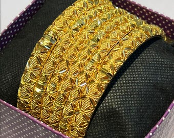 Op de afbeelding: Een set gouden armbanden met een textuur, geometrisch patroon. De armbanden worden gepresenteerd in een paarse en witte polkadot doos met een zwart interieur. De sieraden zijn zeer reflecterend en tonen hun ingewikkelde ontwerp.