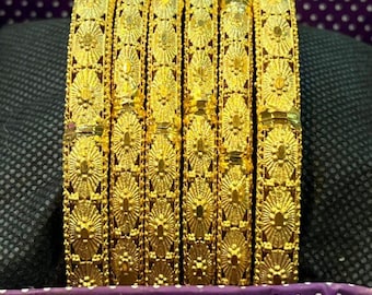 May include: Six gold bangles with intricate floral patterns are displayed in a purple-lined box. The bangles are a bright, shiny gold color and appear to be made of metal. The image includes the text "@umer.jeweller" and "www.umerjeweller.com".
