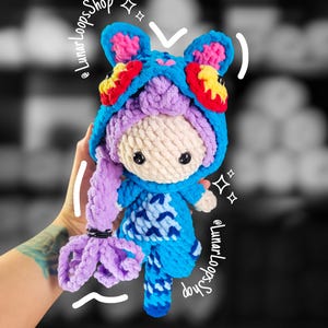 May include: A handmade crochet doll featuring a cream-colored face, black eyes, and a blue hood with red and yellow accents. The doll has long, braided lavender hair and wears a blue outfit with a patterned design. Text reads @LunarLoopsShop.
