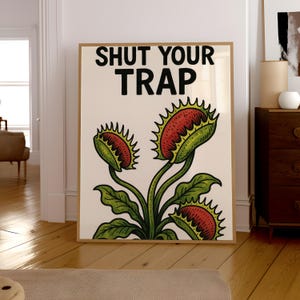 May include: A framed art print featuring a botanical illustration of Venus flytraps. The artwork has a cream-colored background with green leaves and red and green traps. The text "SHUT YOUR TRAP" is printed in bold black letters at the top.