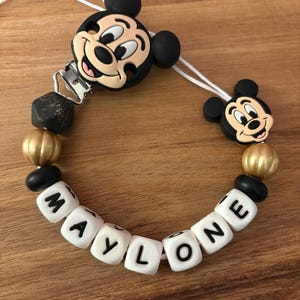 Könnte beinhalten: Ein Schnullerhalter mit zwei schwarzen und beigen Mickey-Maus-Silikonköpfen, goldenen und schwarzen Perlen sowie weißen Würfelperlen, die den Namen MAYLONE in schwarzen Buchstaben bilden. Die Elemente sind auf einer weißen Schnur mit Metallclip.