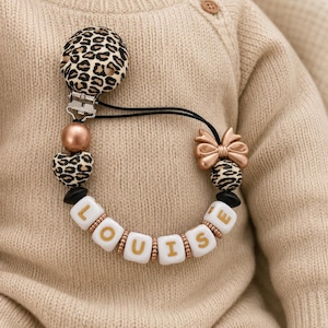 Op de afbeelding: Een speenkoord met een ronde clip met luipaardprint, een metallic roségouden kraal, een hartvormige kraal met luipaardprint, een roségouden strikje en witte vierkante kralen die de naam LOUISE in gouden letters spellen aan een zwart koord.