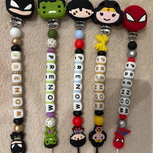 May include: Five pacifier clips featuring superhero-themed silicone character heads including Batman, Hulk, Superman, Wonder Woman, and Spider-Man. Each clip has a string of beads with the word PRENOM spelled out in white lettered cubes.