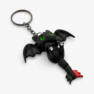 May include: A black dragon keyring with green eyes and white paw pads. The dragon has textured wings and a red tail fin. The keyring is attached to a silver metal ring.