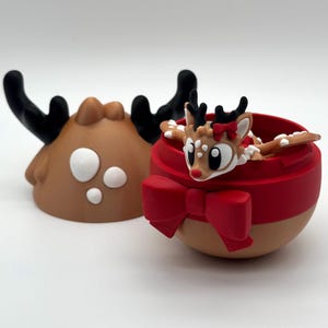 以下が含まれることがあります： 茶色と赤色のクリスマスオーナメント。トナカイのデザイン。オーナメントには赤いリボンと顔を出すトナカイがいます。オーナメントのもう半分は茶色で、黒い角があります。