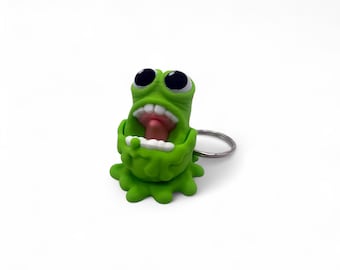 Könnte beinhalten: Ein grüner Monster-Schlüsselanhänger mit großen schwarzen Augen, einem weit geöffneten Mund und einer rosa Zunge. Der Schlüsselanhänger besteht aus einem weichen Material und hat einen silbernen Metallring. Das Monster hat ein cartoonartiges Design.