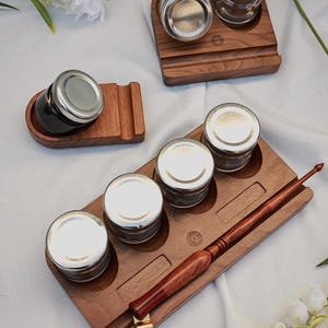 Könnte beinhalten: Holz-Tintenfass-Sets mit Gläsern und silbernen Deckeln. Die Sets umfassen Holztabletts und einen Stifthalter. Ein Kalligraphiestift aus Holz mit goldener Feder ist ebenfalls enthalten. Das Holz hat eine satte, dunkelbraune Farbe.