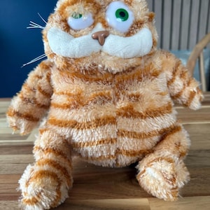 Könnte beinhalten: Ein Stofftier in Form einer orange-braun gestreiften Katze mit großen, ausdrucksstarken Augen und einer weißen Schnauze. Die Katze hat eine braune Nase und weiße Schnurrhaare. Es liegt auf einer Holzoberfläche.