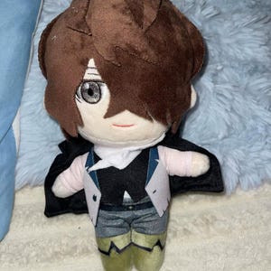 Puede incluir: Una muñeca de peluche con cabello castaño y un rostro detallado, que lleva un chaleco negro, una camisa blanca y pantalones grises. La muñeca tiene una capa negra y botas verdes. La muñeca está sobre una superficie azul claro.