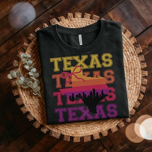 May include: Black t-shirt with the word "TEXAS" in yellow, orange, and purple. The shirt features a Texas-shaped graphic with a sunset design and a city skyline silhouette. The shirt is folded on a woven mat.