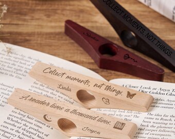May include: Wooden book page holders with engraved text, including "Collect moments, not things." and "A reader lives a thousand lives." One is dark brown, one is burgundy, and two are light wood. The holders are resting on open books.