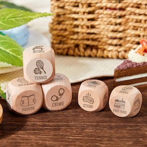 Può includere: Dadi di legno con icone e testo incisi, tra cui "Tennis", "Lettura" e "Steak". È visibile anche una fetta di torta al cioccolato con panna e fragole. I dadi sono su una superficie di legno.