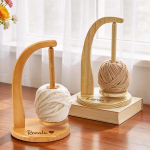 May include: Two wooden yarn holders with arched supports and central dowels. One holds a white yarn ball, the other a tan yarn ball. The bases are engraved with names. A vase of flowers sits nearby.