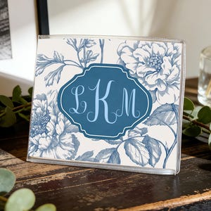Op de afbeelding: Een heldere acryl fotolijst met een blauw en wit bloemenpatroon. De lijst heeft een donkerblauw label met de letters "LKM" in lichtblauw schrift. Het staat op een donker houten oppervlak.