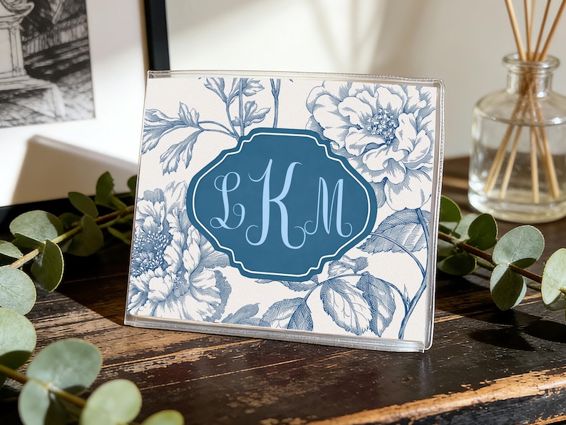 Op de afbeelding: Een heldere acryl fotolijst met een blauw en wit bloemenpatroon. De lijst heeft een donkerblauw label met de letters "LKM" in lichtblauw schrift. Het staat op een donker houten oppervlak.