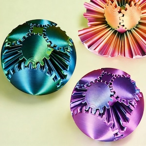 May include: Three decorative spheres with gear-shaped designs in shades of blue-green, pink-orange, and purple. The objects are set against a pale yellow background. The spheres have a metallic sheen and intricate details.