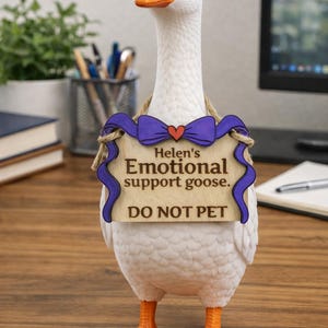 May include: A white goose figurine with an orange beak and feet, wearing a wooden sign suspended by twine. The sign features a purple bow with a red heart and the text: Helen's Emotional support goose. DO NOT PET. The goose stands upright.